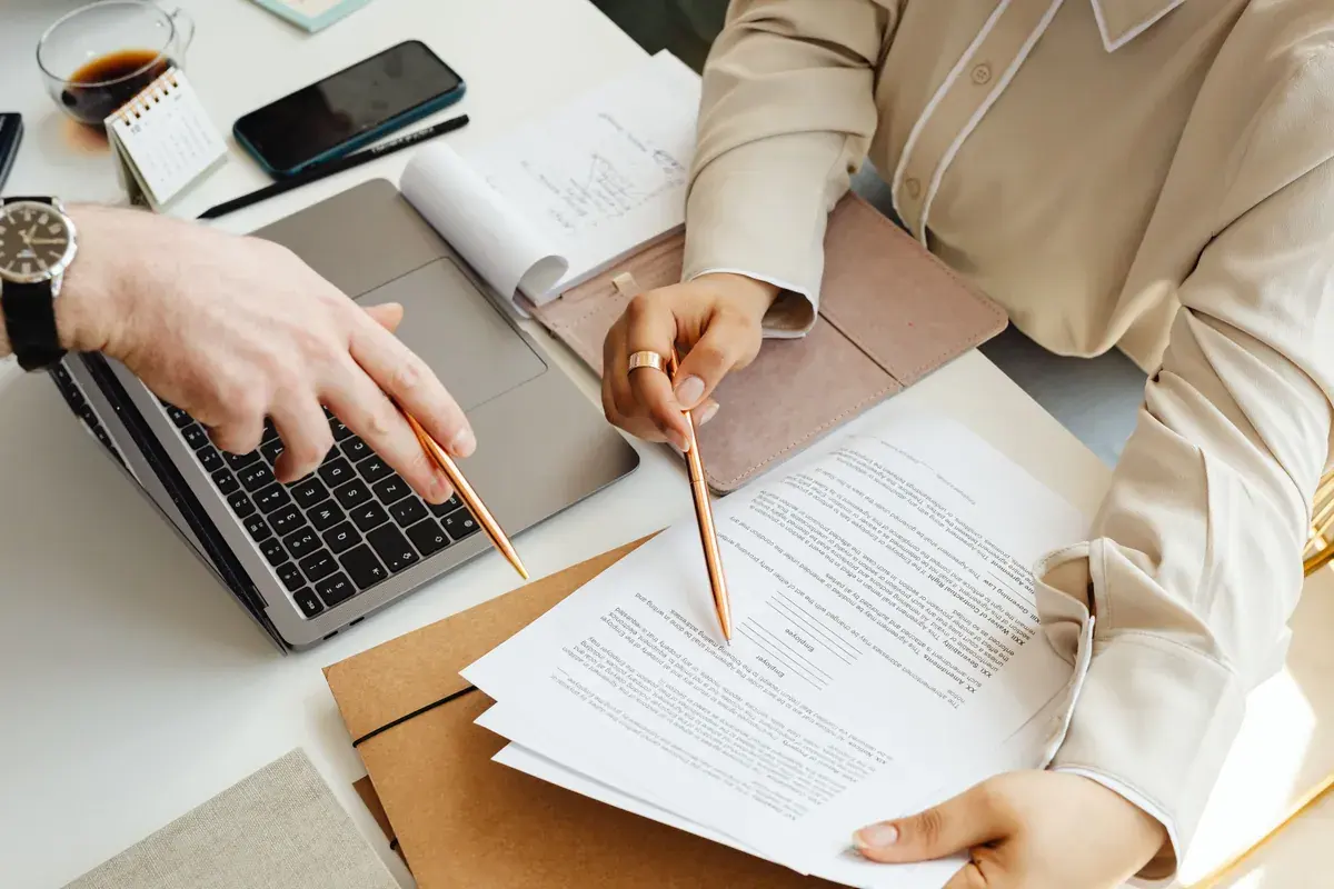 Two people at a shared workspace pointing pens at a stack of printed documents next to a laptop