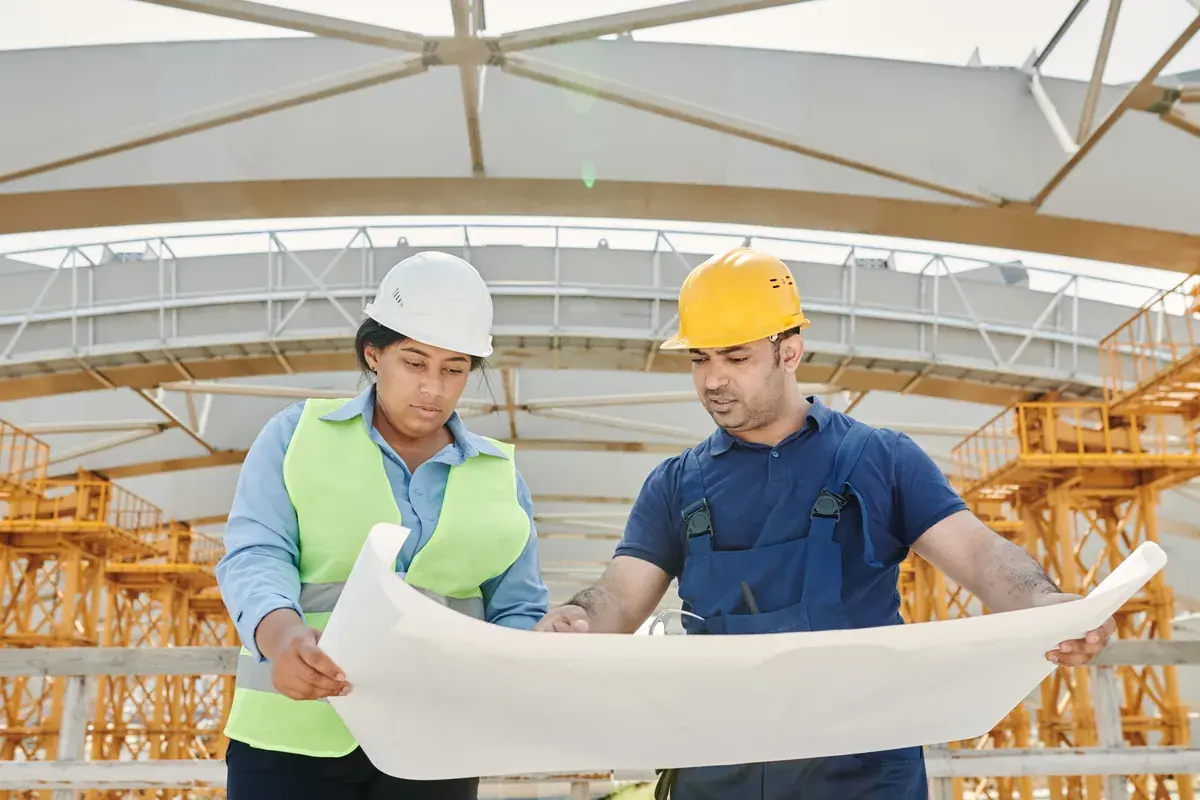 Two construction professionals in safety vests and hard hats reviewing a set of large printed plans on a job site