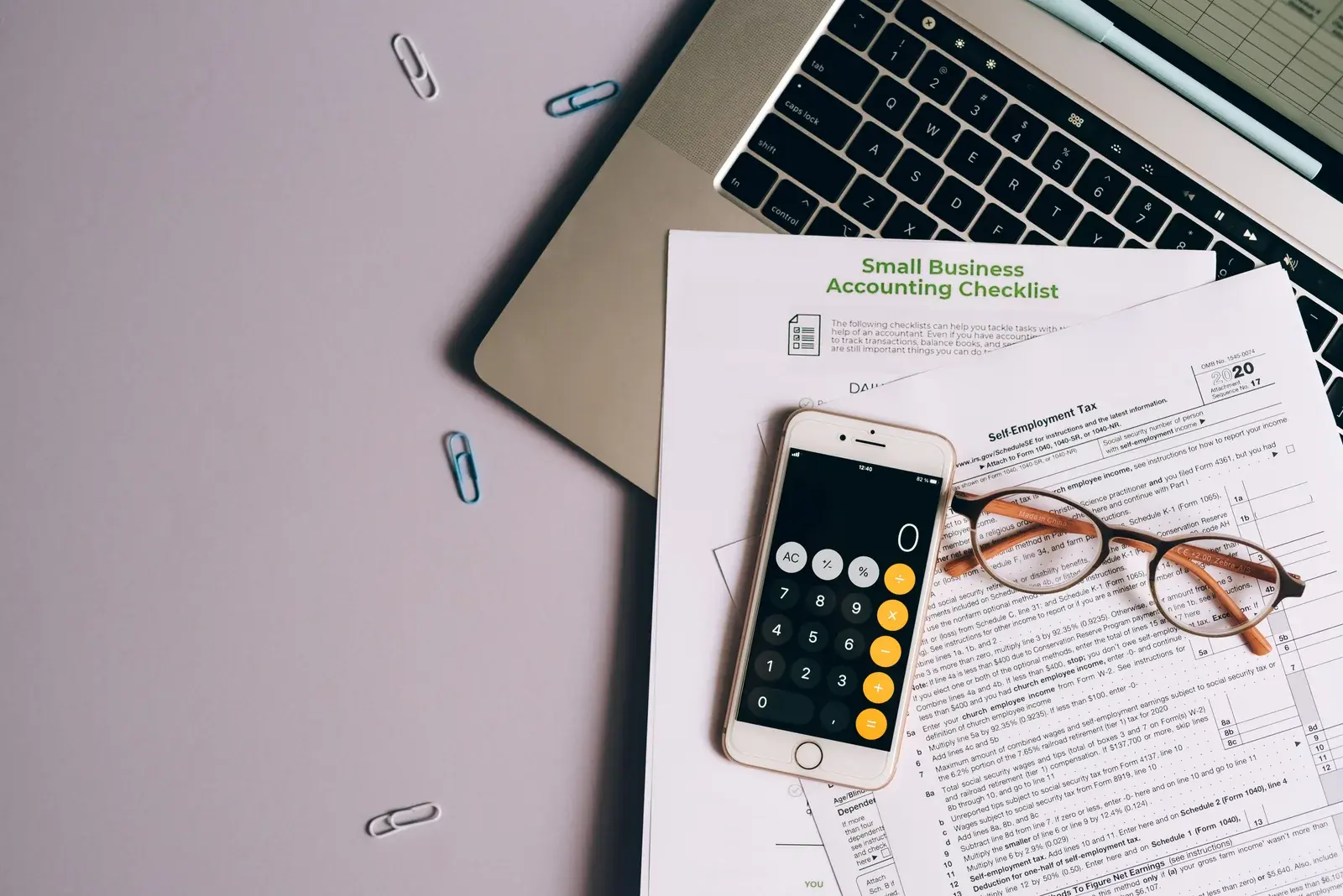 A workspace with a printed accounting checklist next to a smartphone calculator, reading glasses, and a laptop