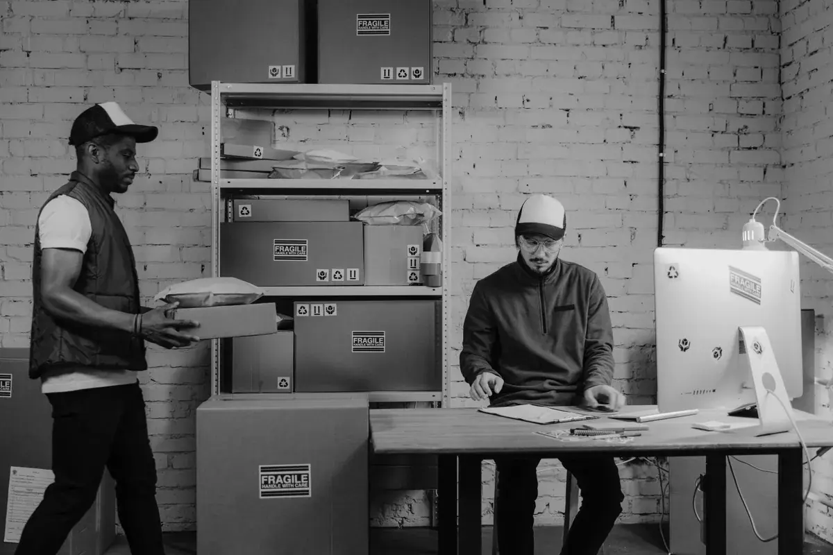 Two workers in a small fulfillment room move labeled shipping boxes and review orders at a desk.