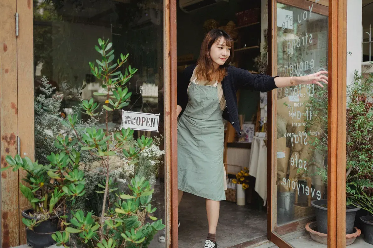 A shop owner in an apron opens the glass door of a small plant filled storefront.