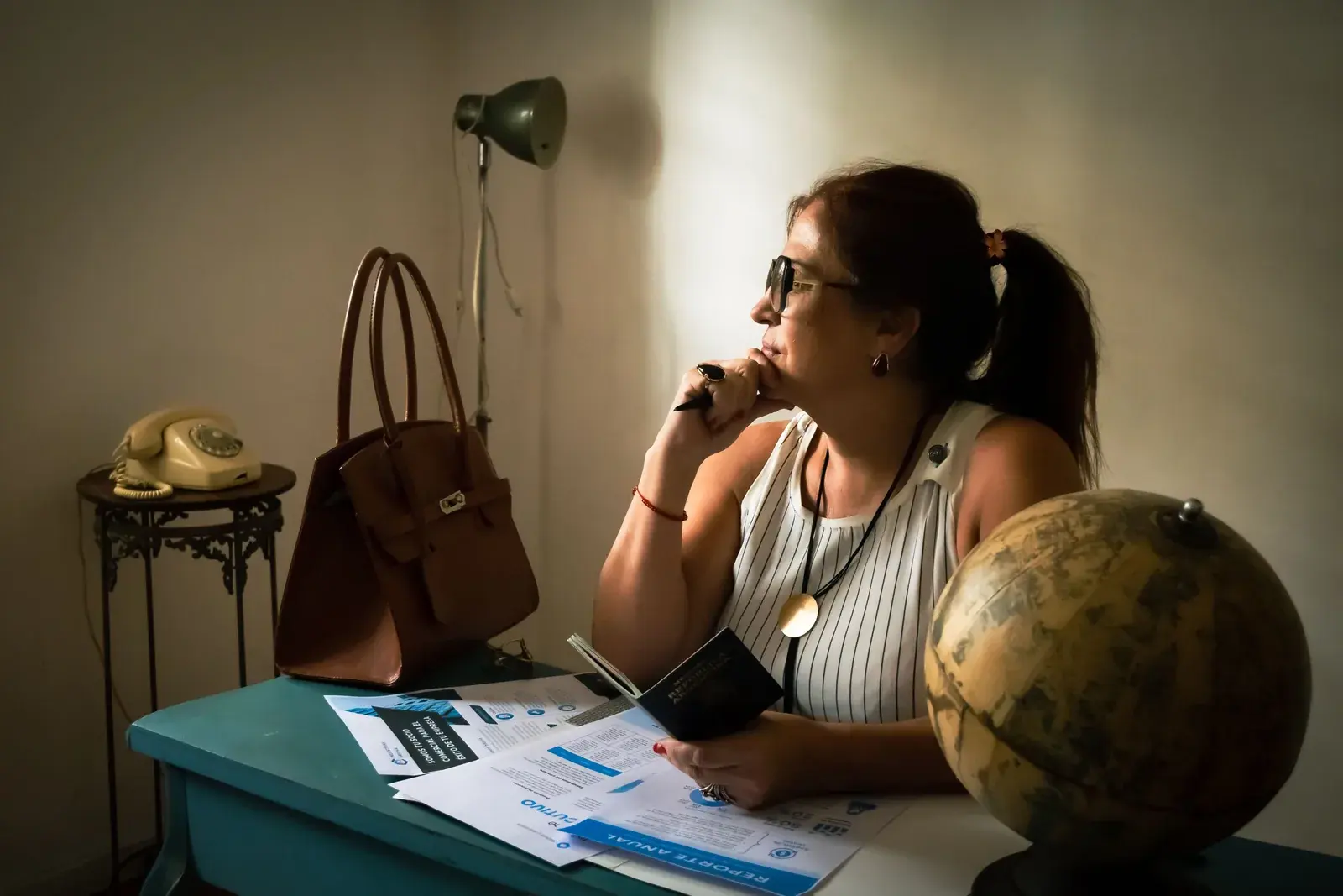 A woman pauses at a desk with a globe, a passport, and printed filings beside her.