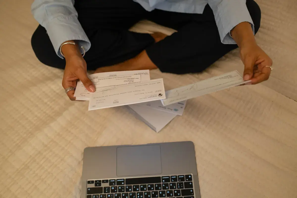 Two hands holding printed checks beside an open laptop on a light desk