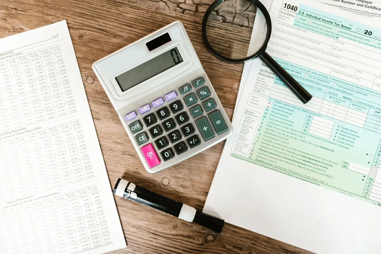 An overhead view of printed tax forms, a calculator, and a magnifying glass arranged on a wooden desk for review