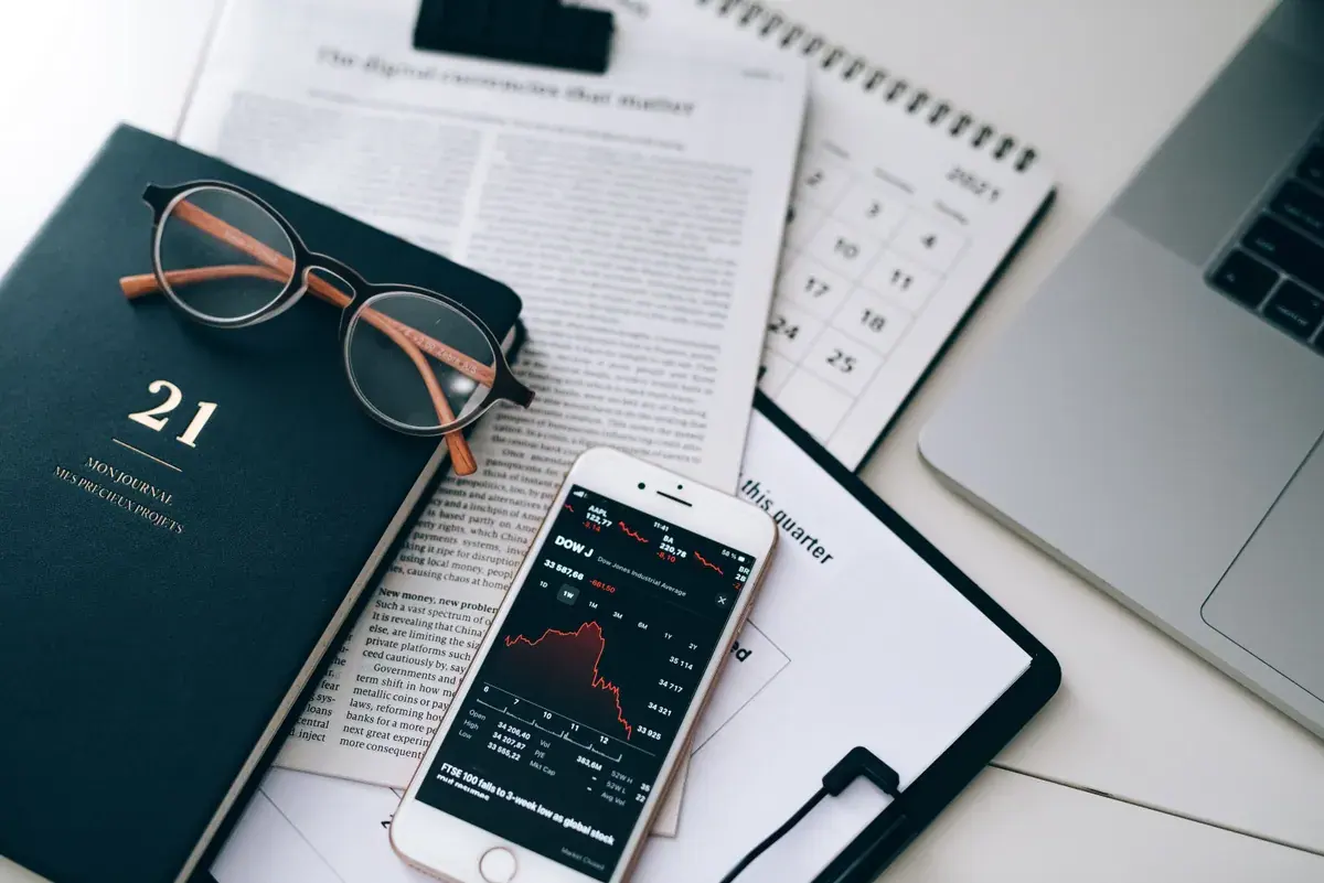 Flat lay of a dark journal, reading glasses, a newspaper and a phone showing a market chart.