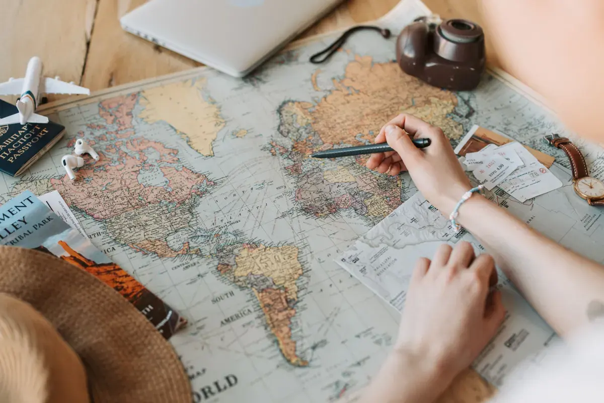 A pair of hands marking destinations on a world map with a passport and a camera on the desk