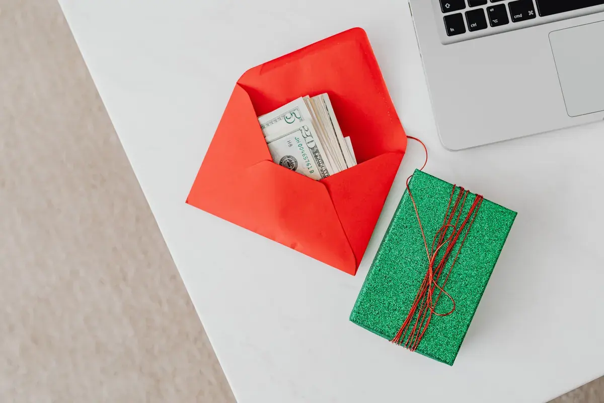 An open envelope of cash beside a wrapped gift box on a white desk next to a laptop
