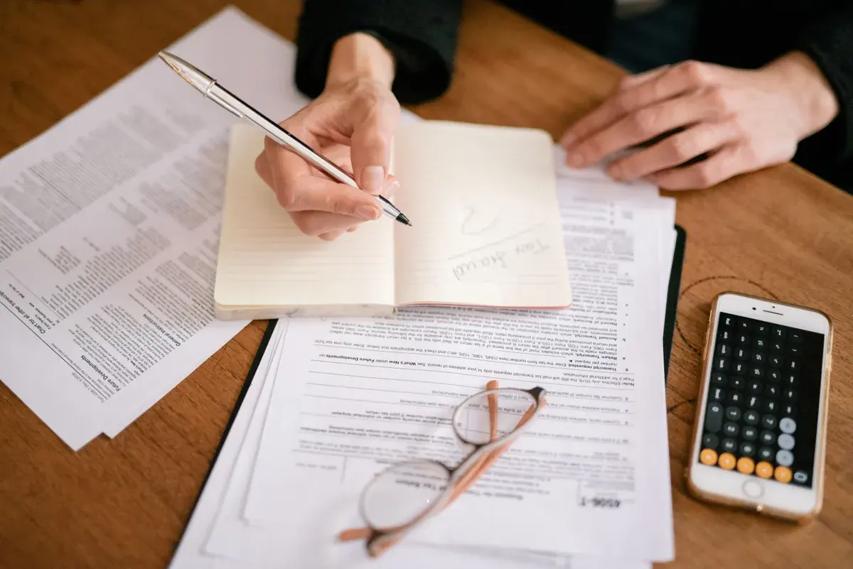 Hands writing notes in an open notebook resting on printed tax forms beside a calculator