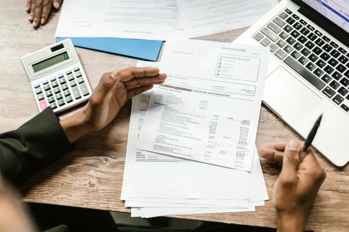 Hands hold a printed account statement above a wooden desk with a calculator and a laptop.