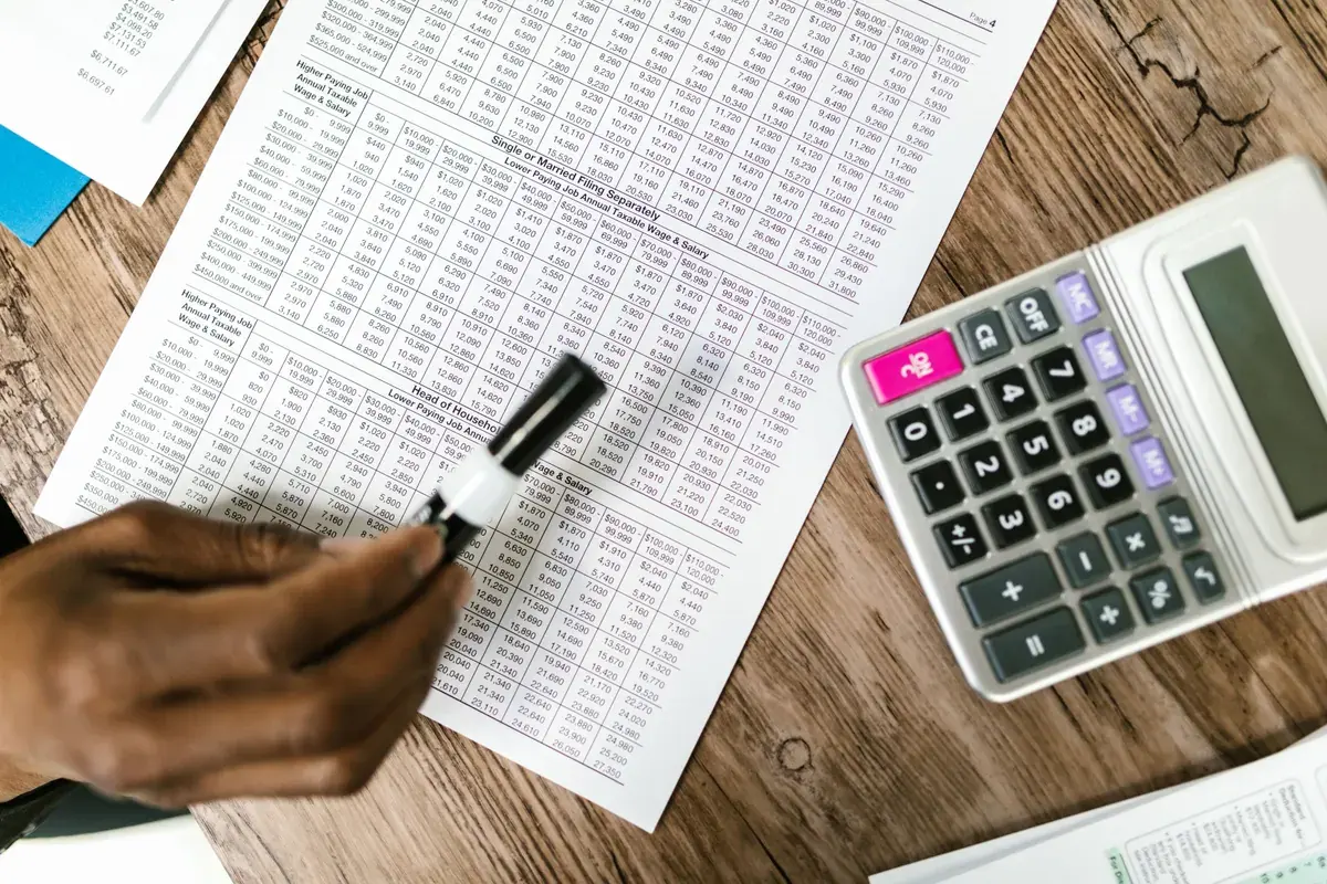 A hand holds a marker over printed tax tables next to a calculator on a wooden surface.