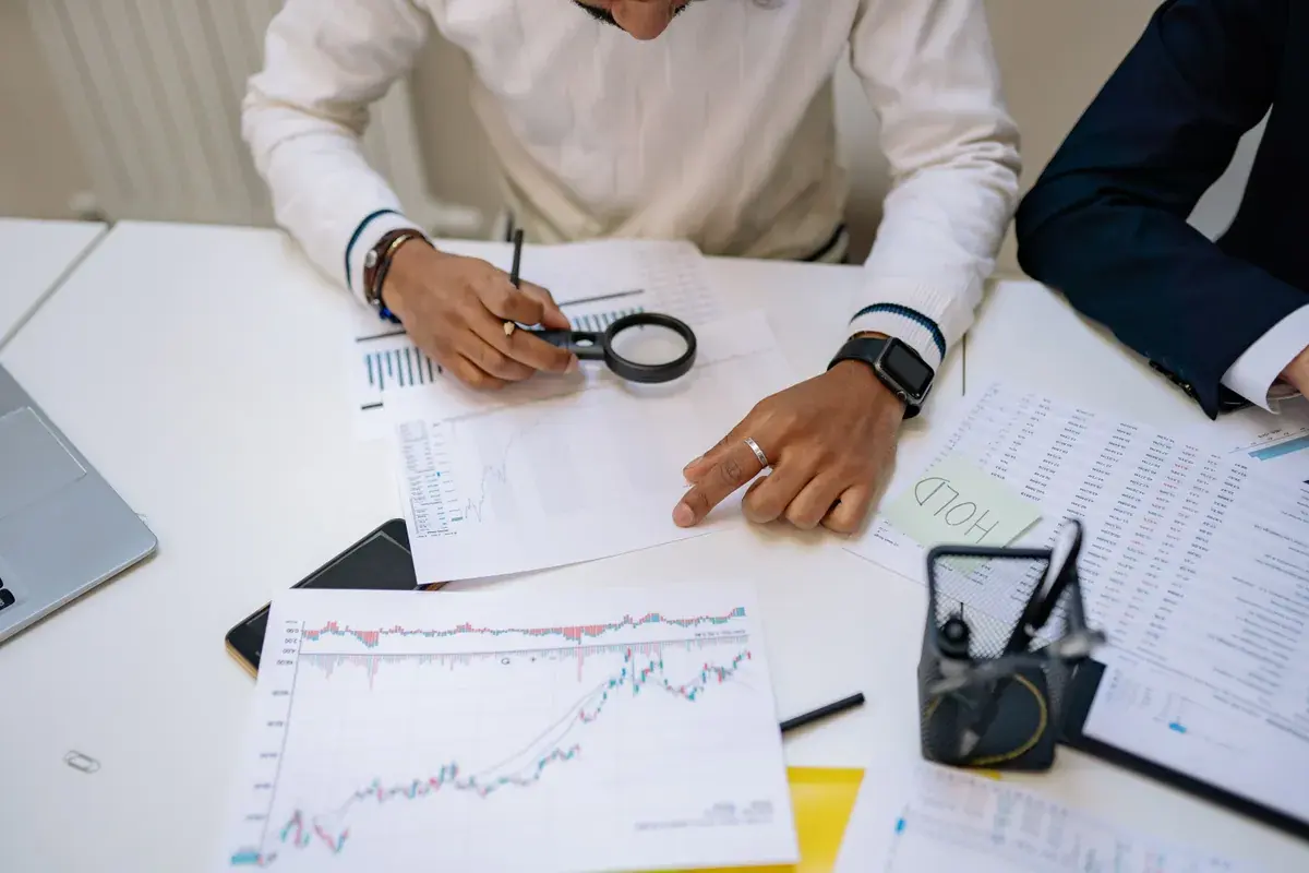 Two business professionals review printed stock charts with a magnifying glass at a conference table