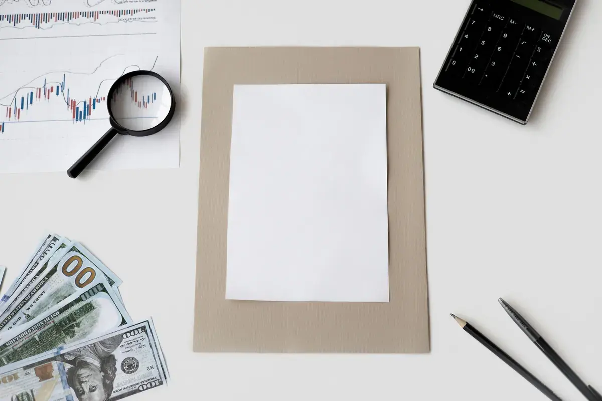 Flat lay of a blank sheet, a calculator, printed stock charts and a magnifying glass on a light desk