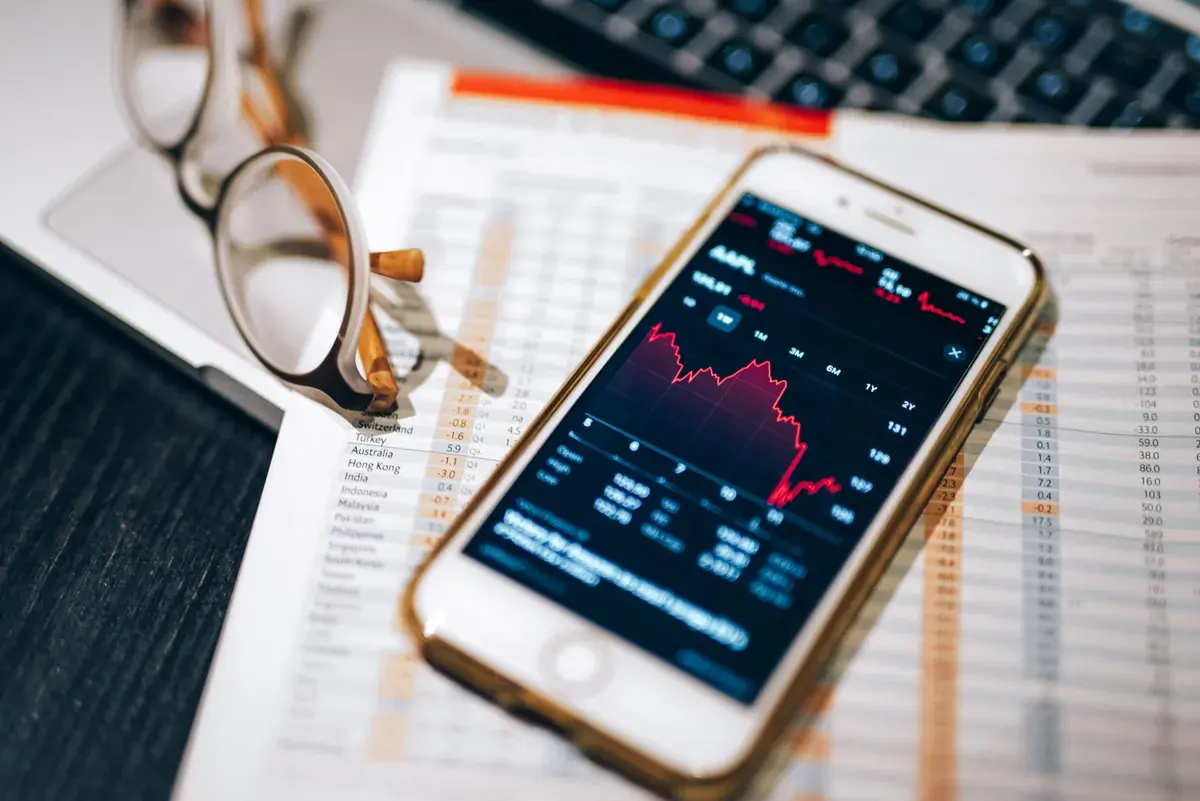 A phone displaying a stock chart rests on a printed financial table beside a pair of eyeglasses