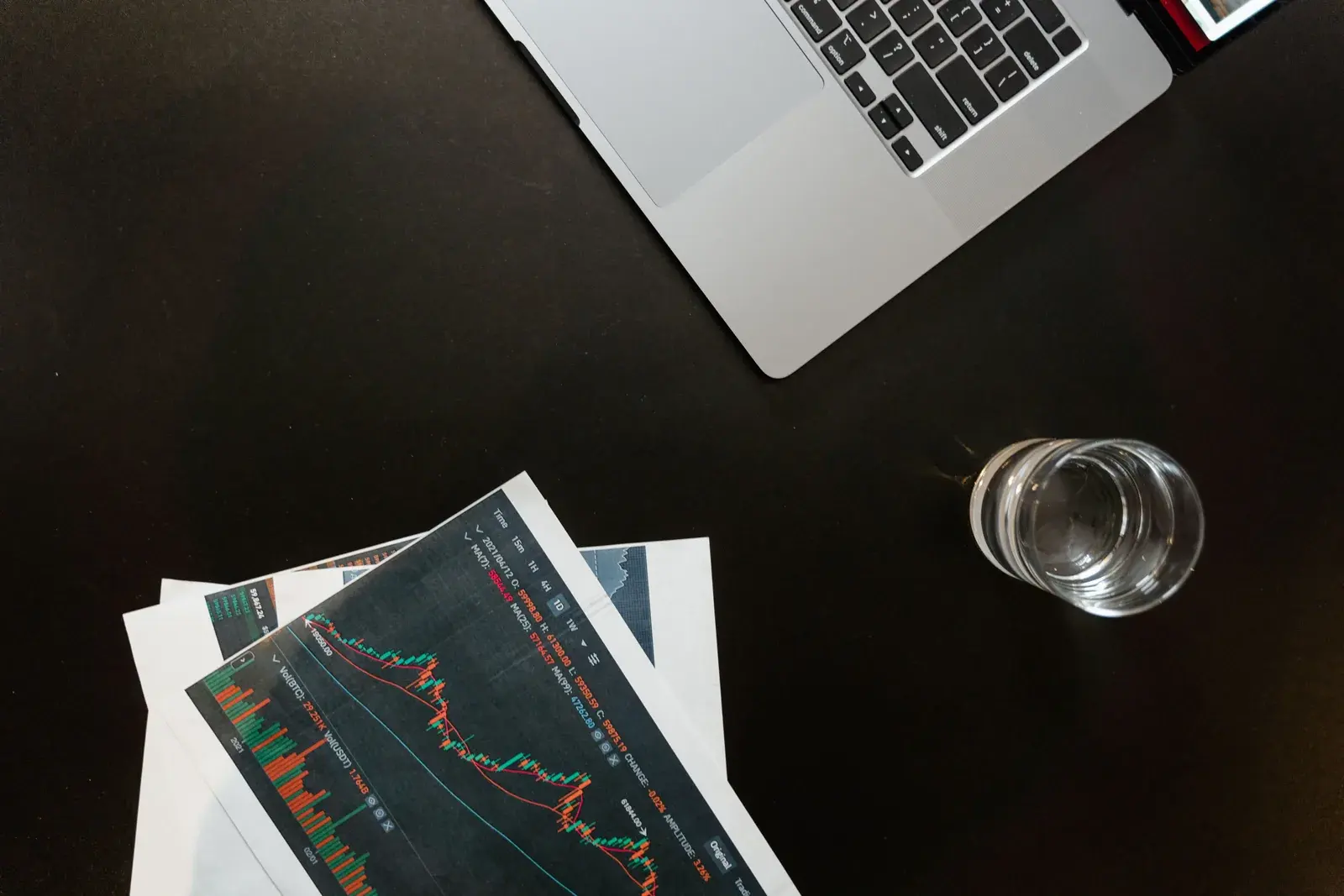 A silver laptop sits on a dark desk with printed stock charts and a glass of water nearby