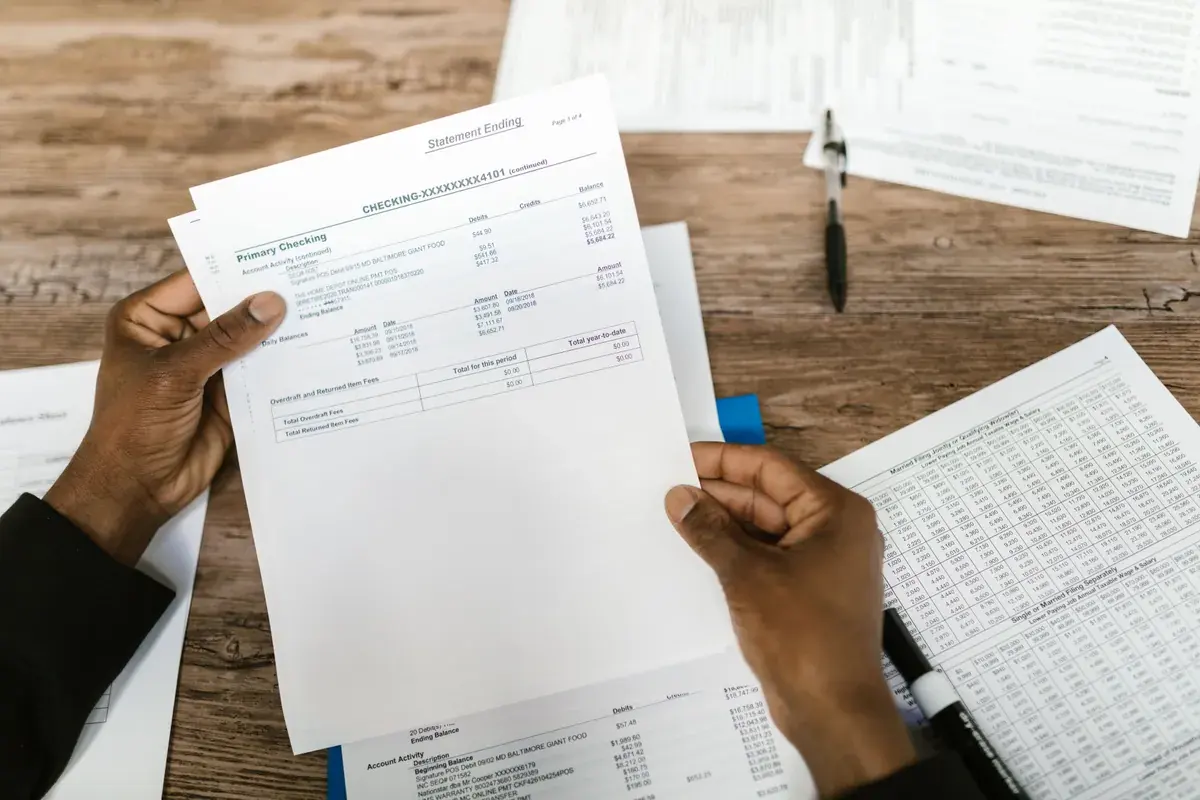 Hands holding a printed checking account statement over a wooden desk alongside other paperwork