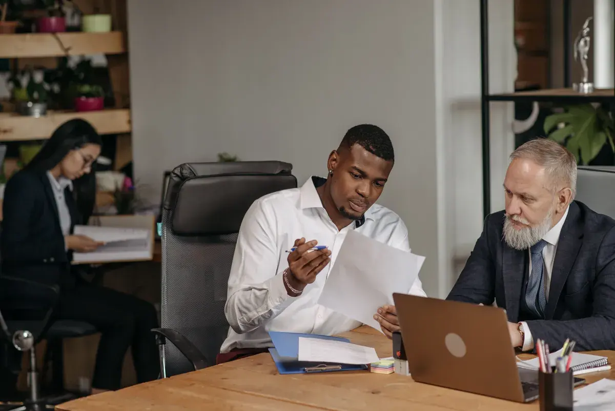 Two professionals examining a printed document together at a shared desk with a laptop in a bright office