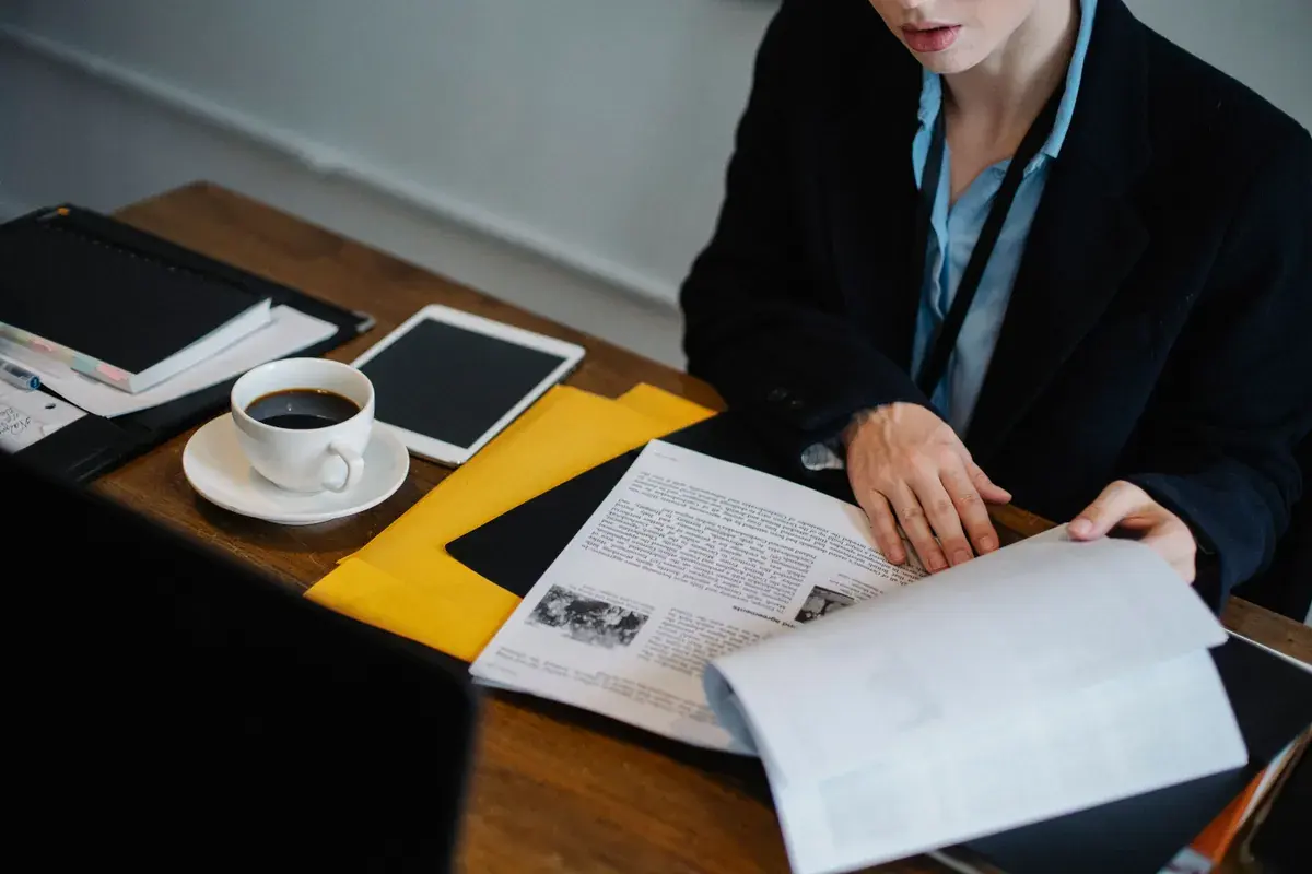 A person at a dark wooden desk turning the pages of a printed report beside a tablet