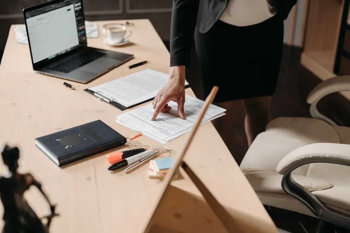 A printed form being completed on a wooden desk beside a laptop and a notebook