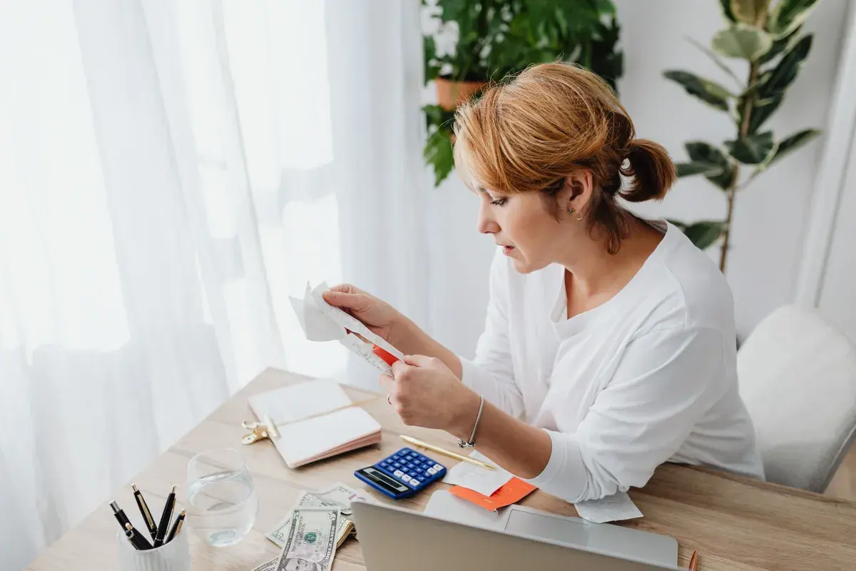 A person at a sunlit desk reads a paper receipt with a notebook, calculator and laptop in front of them.