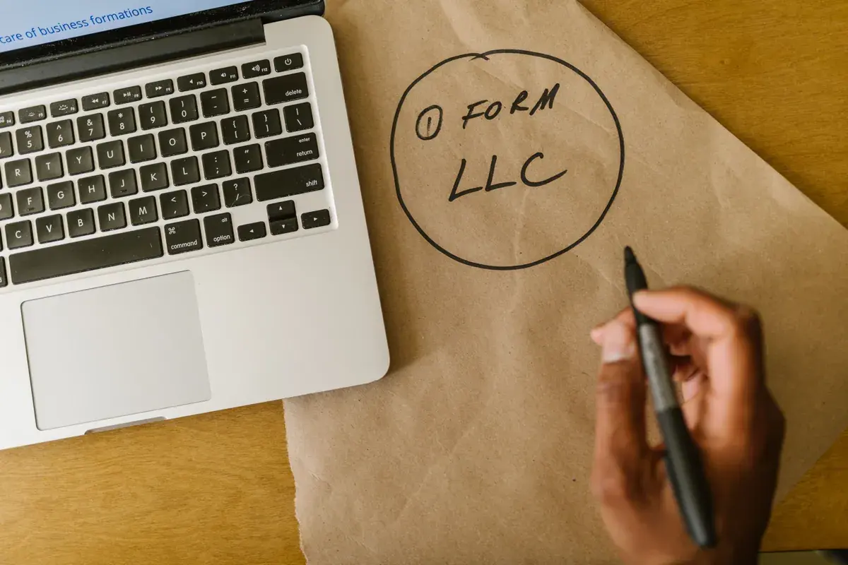 A hand writes a short note about forming an LLC on brown paper beside an open laptop.