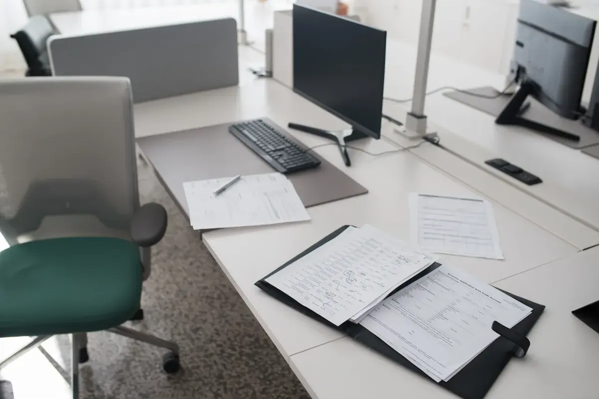 Un escritorio de oficina con monitor, teclado y formularios impresos en blanco abiertos en una carpeta junto a una silla vacía.