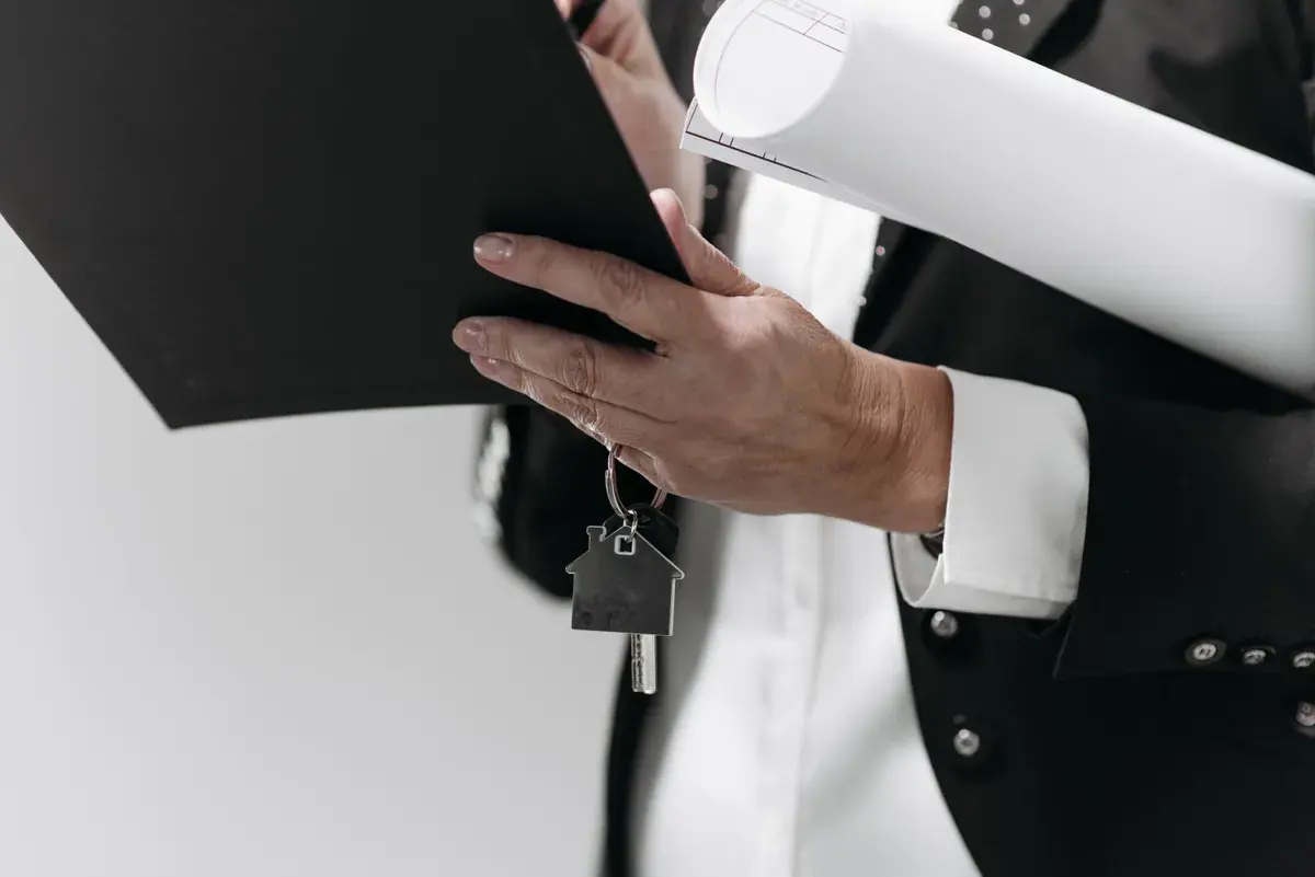 A professional in a dark suit holds a folder with a small house key attached beside rolled architectural drawings