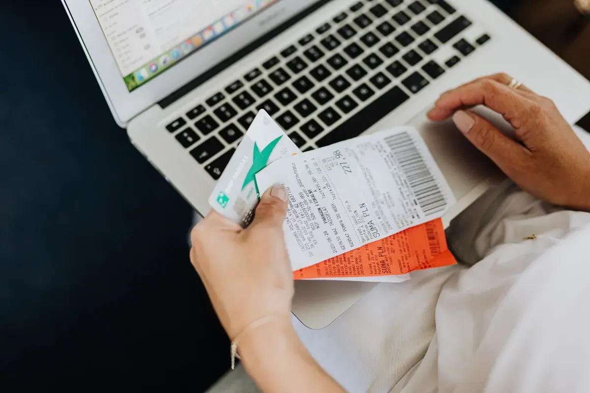 A close-up of hands holding paper receipts and a plain bank card above an open laptop keyboard