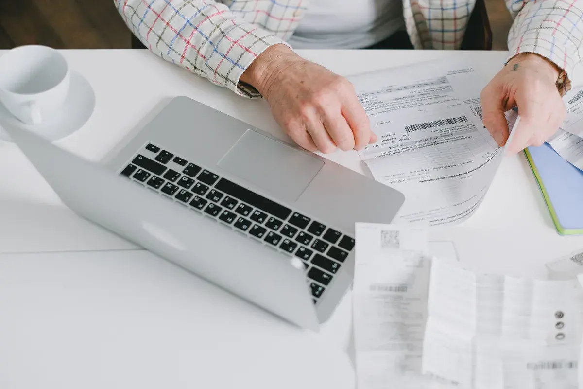 Close-up of hands organizing printed application forms next to a laptop on a plain desk