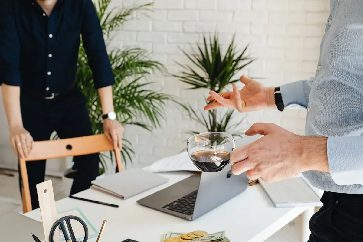 Two colleagues stand across a bright office desk with an open laptop, notebooks and a glass mug of coffee