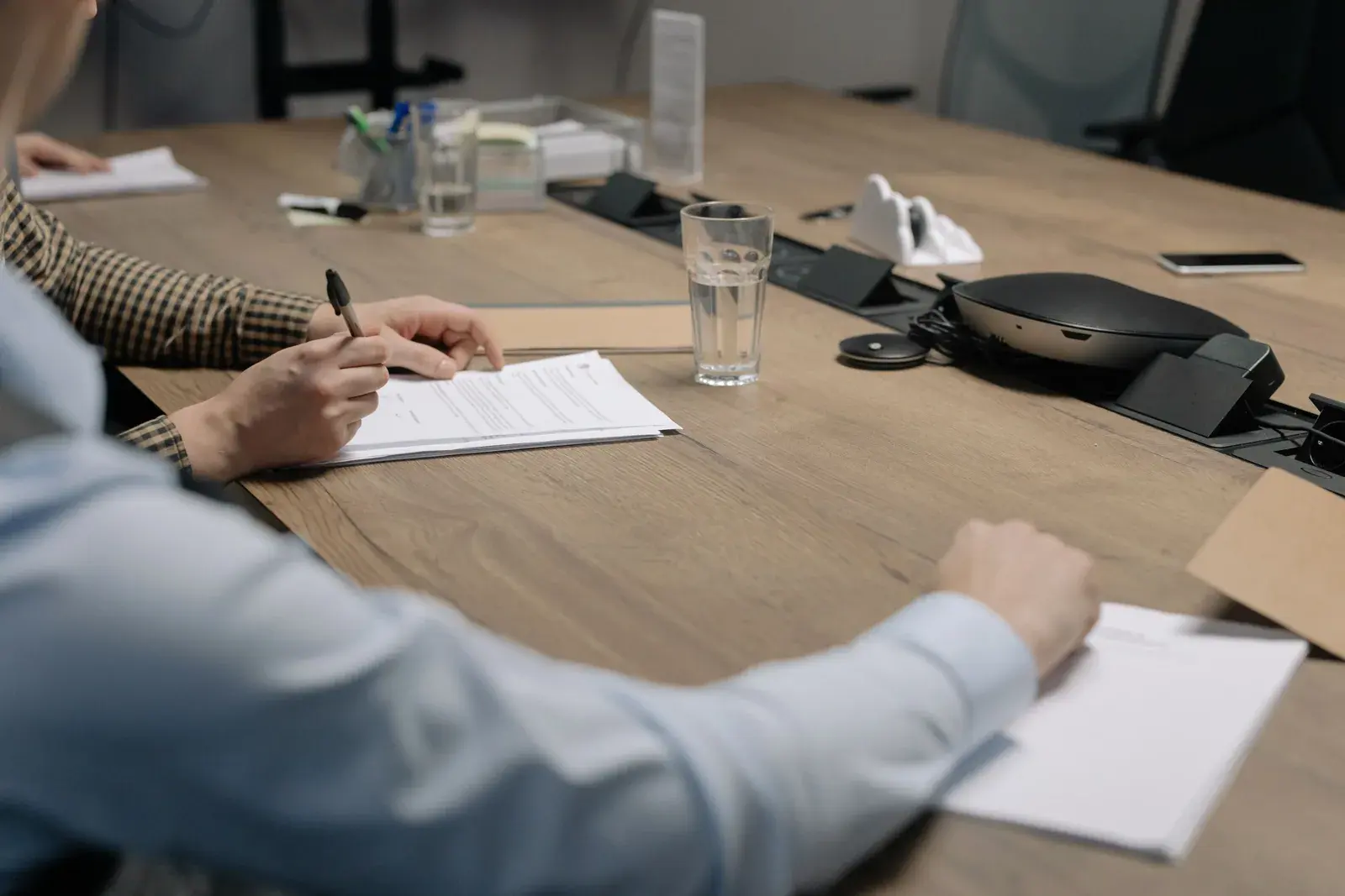 Two people at a wood conference table review and sign a printed document with pens in hand