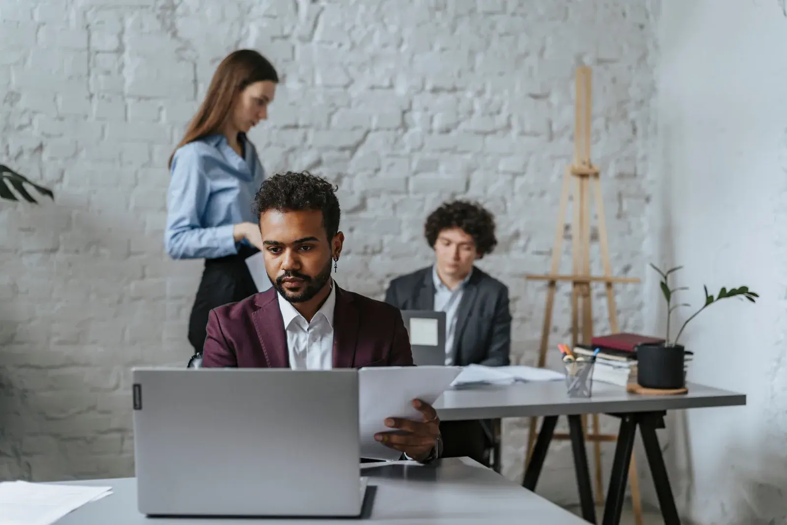 A professional in a burgundy blazer reviews a document at a laptop, with two colleagues working in the background