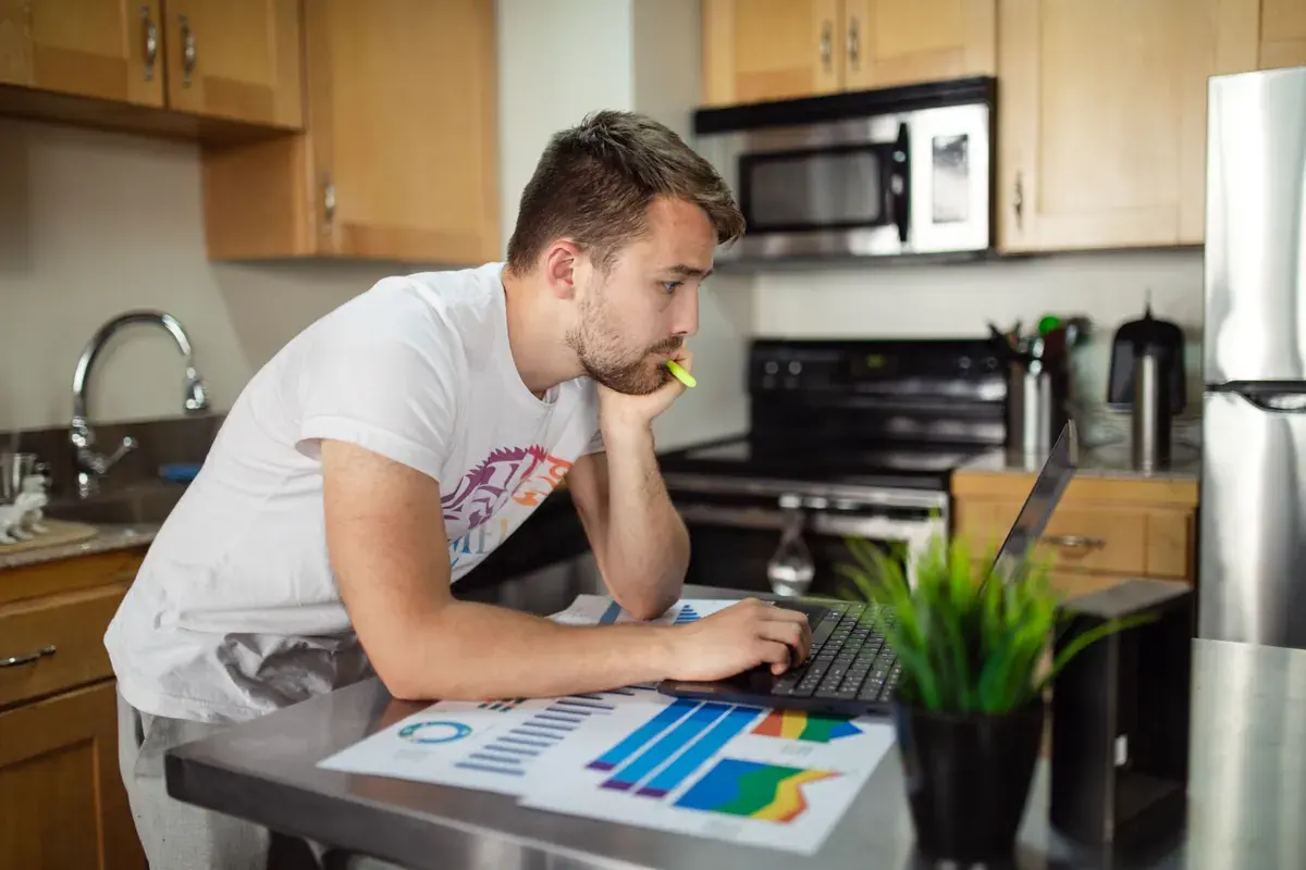 Un hombre trabaja solo en una computadora portatil sobre la barra de la cocina con graficos impresos a su lado