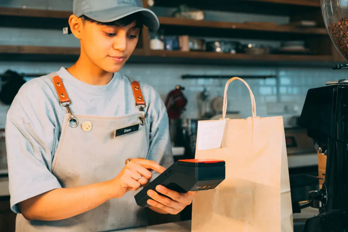 A cafe worker in an apron taps a handheld card reader beside a paper bag on the counter