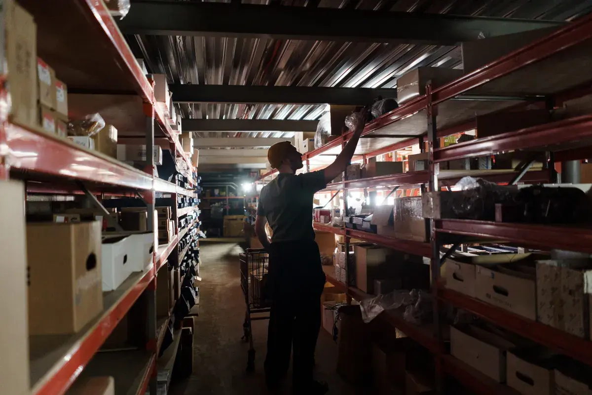 A worker reaches for stock on a tall shelf in a dim warehouse aisle lined with boxes