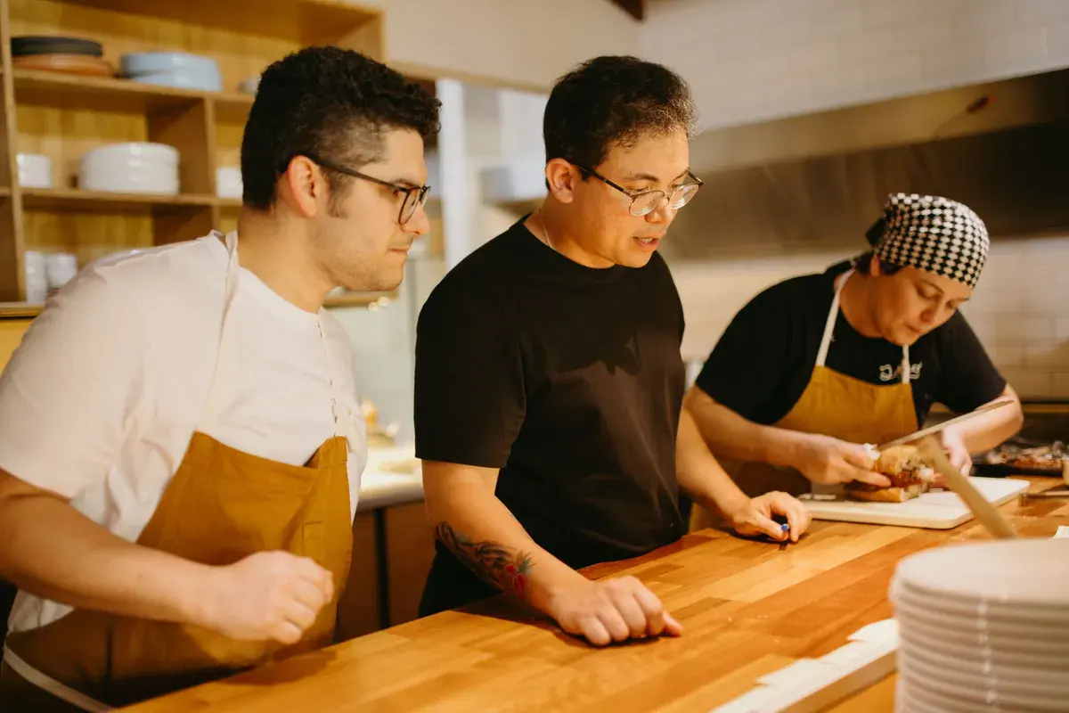 Tres trabajadores de restaurante con delantales preparan comida juntos en un mostrador de cocina de madera