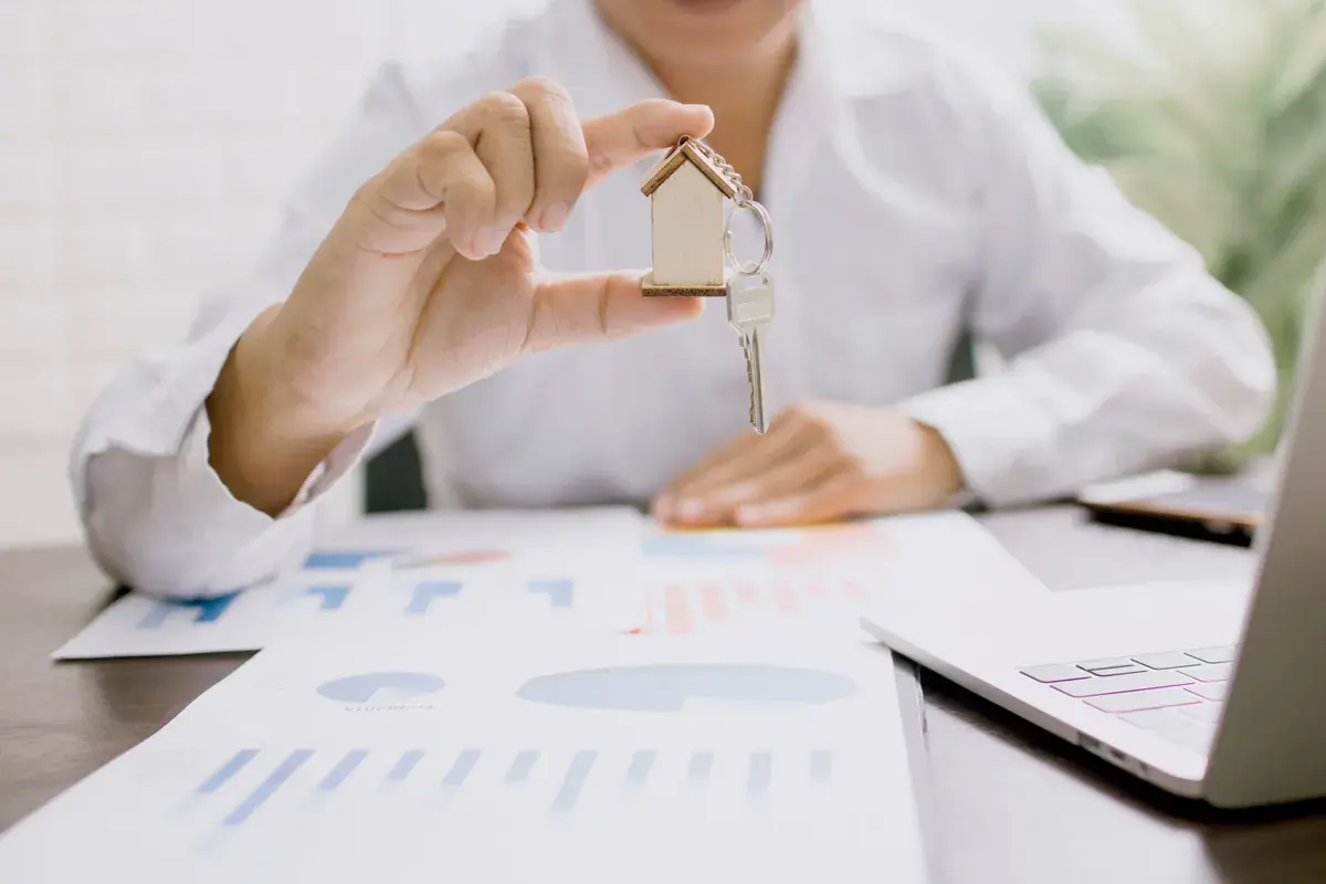 Hands holding a small house model and a key above printed property charts