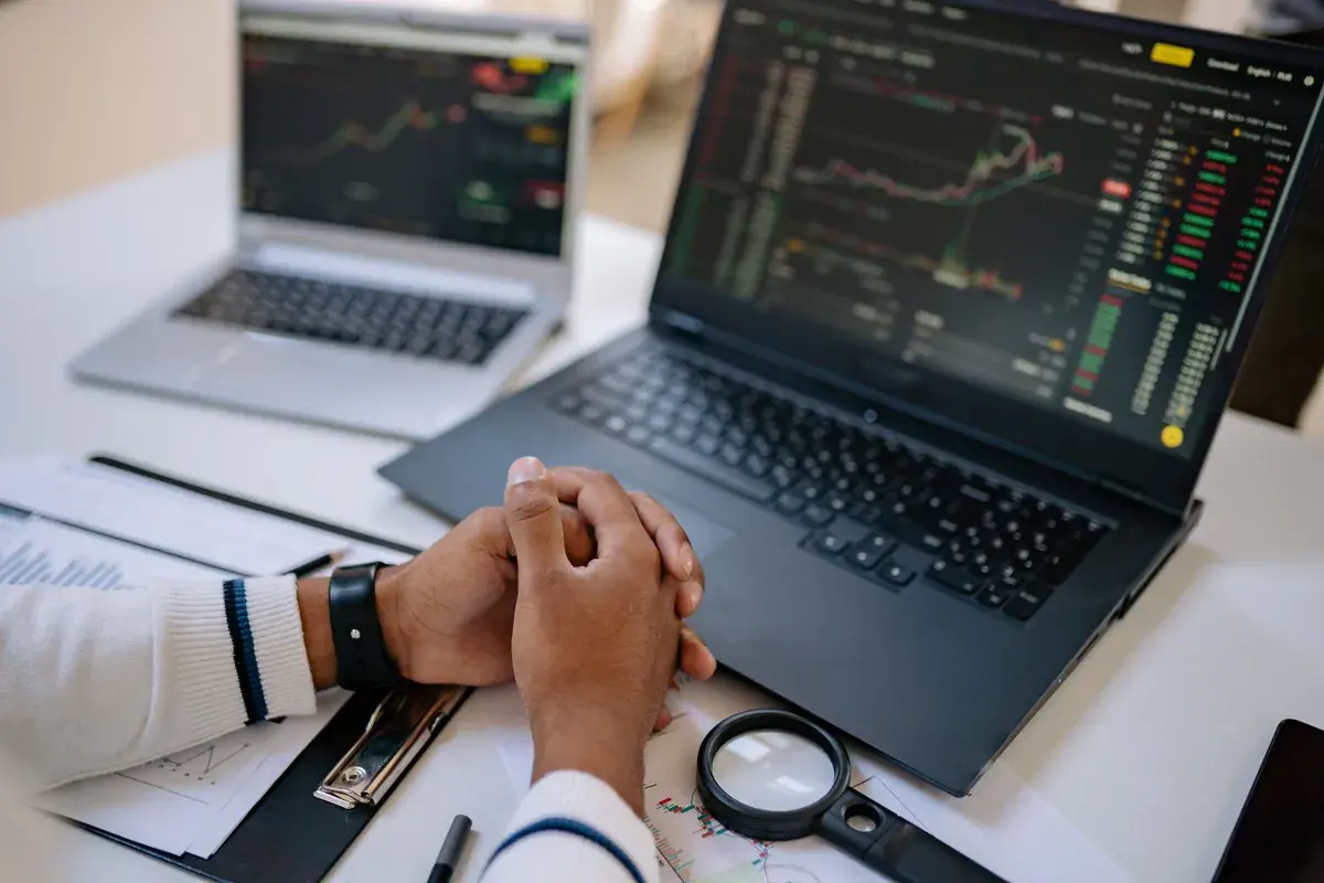 Folded hands rest beside an open laptop showing market charts and a magnifying glass