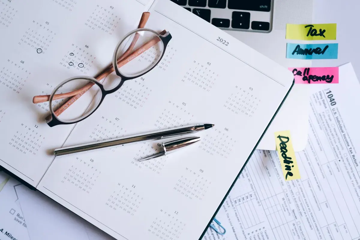An open planner with reading glasses and a pen beside a printed tax form and sticky notes
