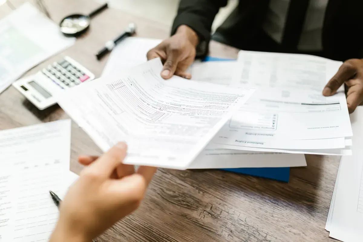 Two people passing printed tax forms across a wooden desk with a calculator