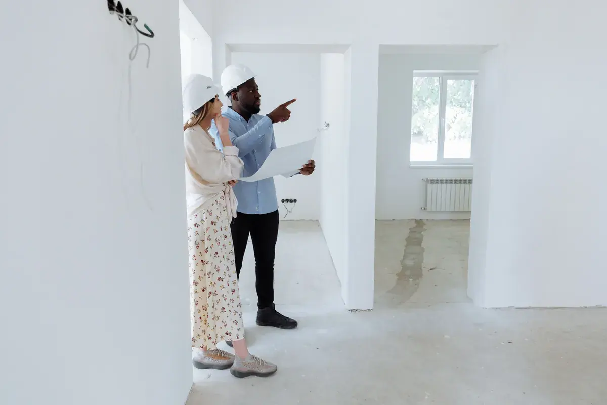 Two people in hard hats reviewing a printed building plan inside an empty white room