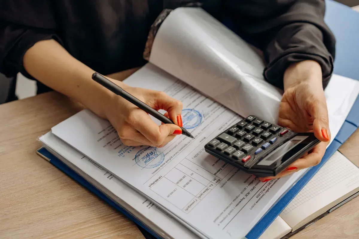 Hands using a calculator while signing stamped financial documents at a desk.