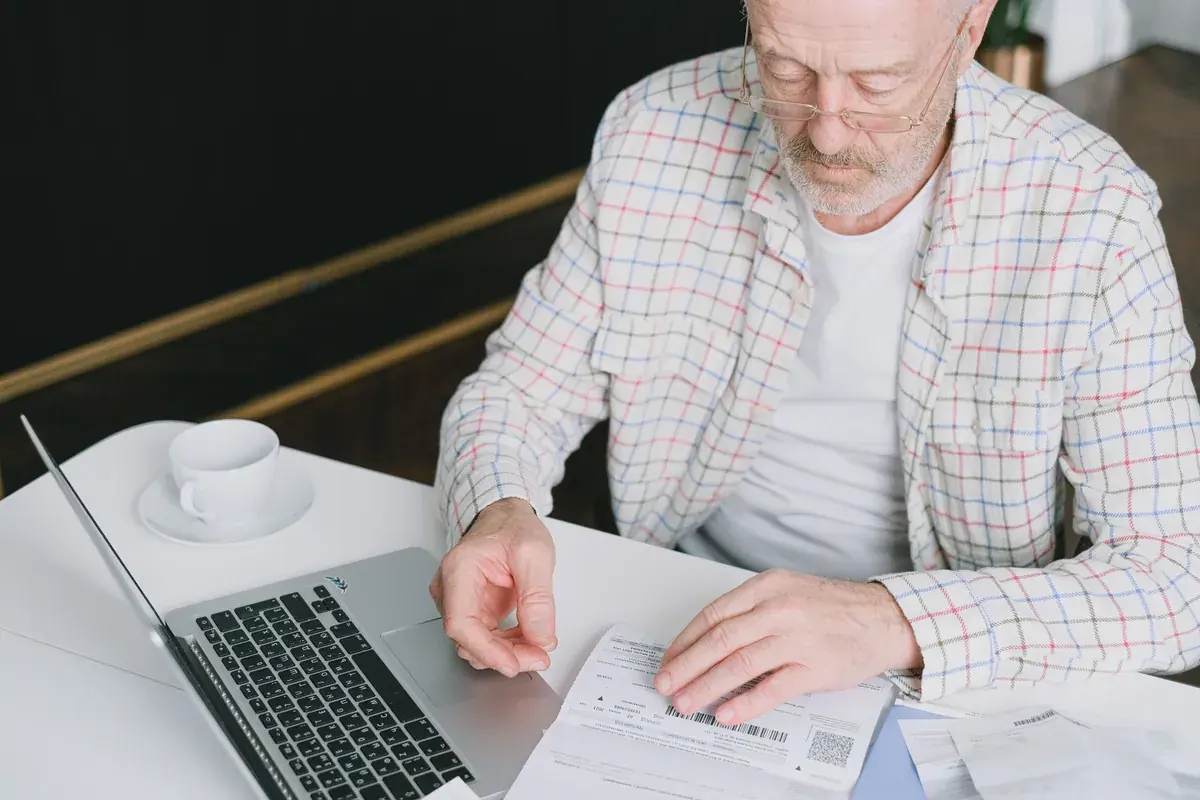 Un hombre mayor con lentes revisa estados impresos junto a una laptop en una mesa blanca