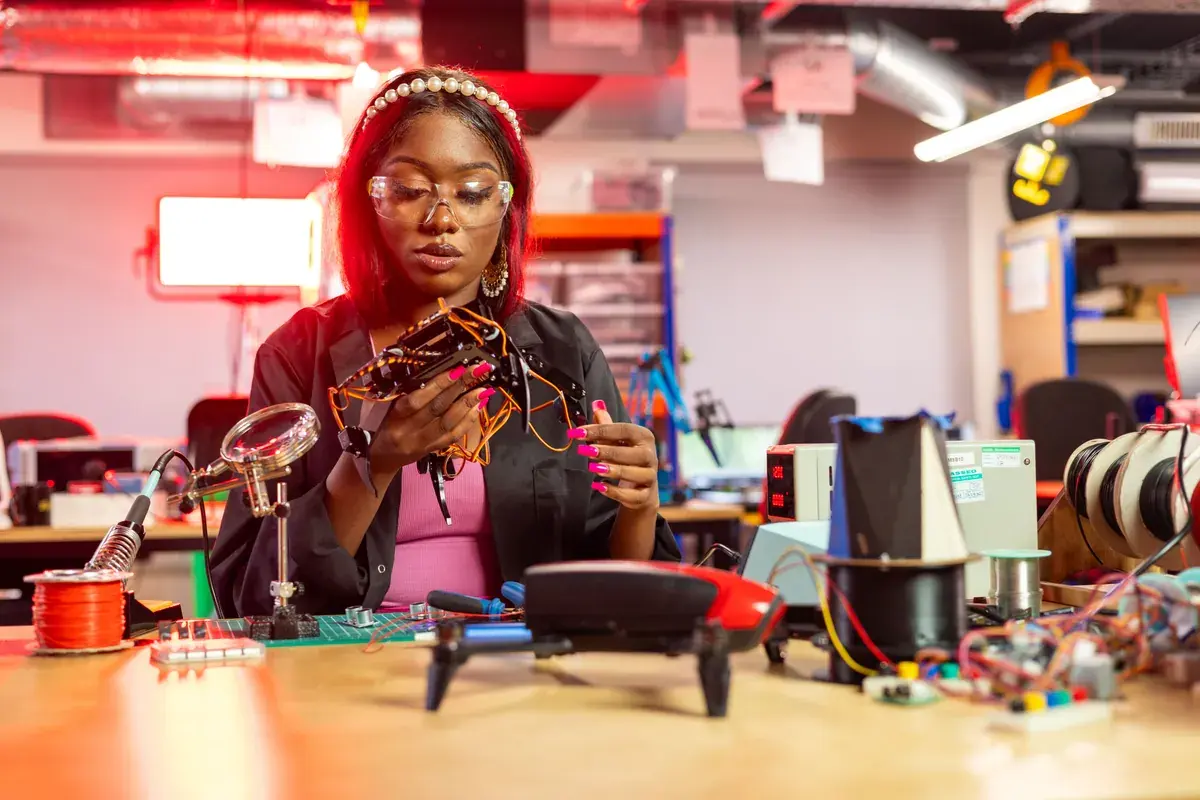 A person in safety glasses inspects a small circuit assembly at a workbench full of tools and parts.