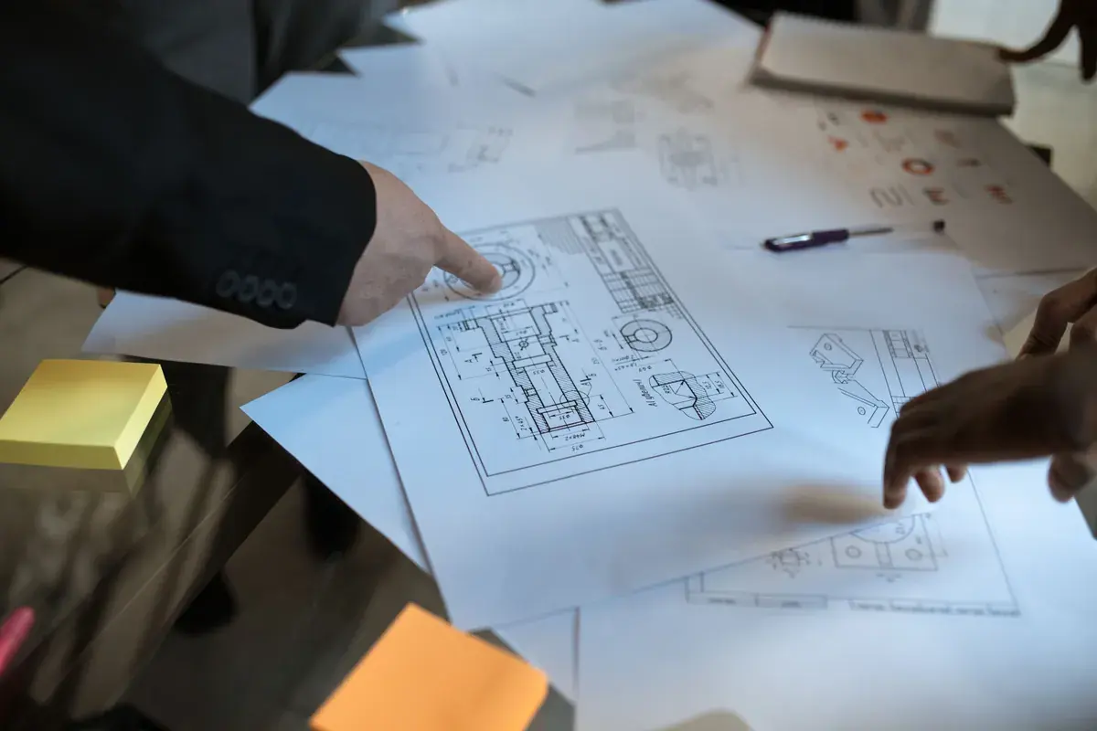 Several hands point at mechanical part drawings spread across a dark desk with sticky notes and a pen.