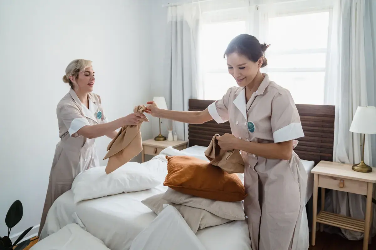 Two housekeepers in uniform making a bed with fresh linens in a bright room