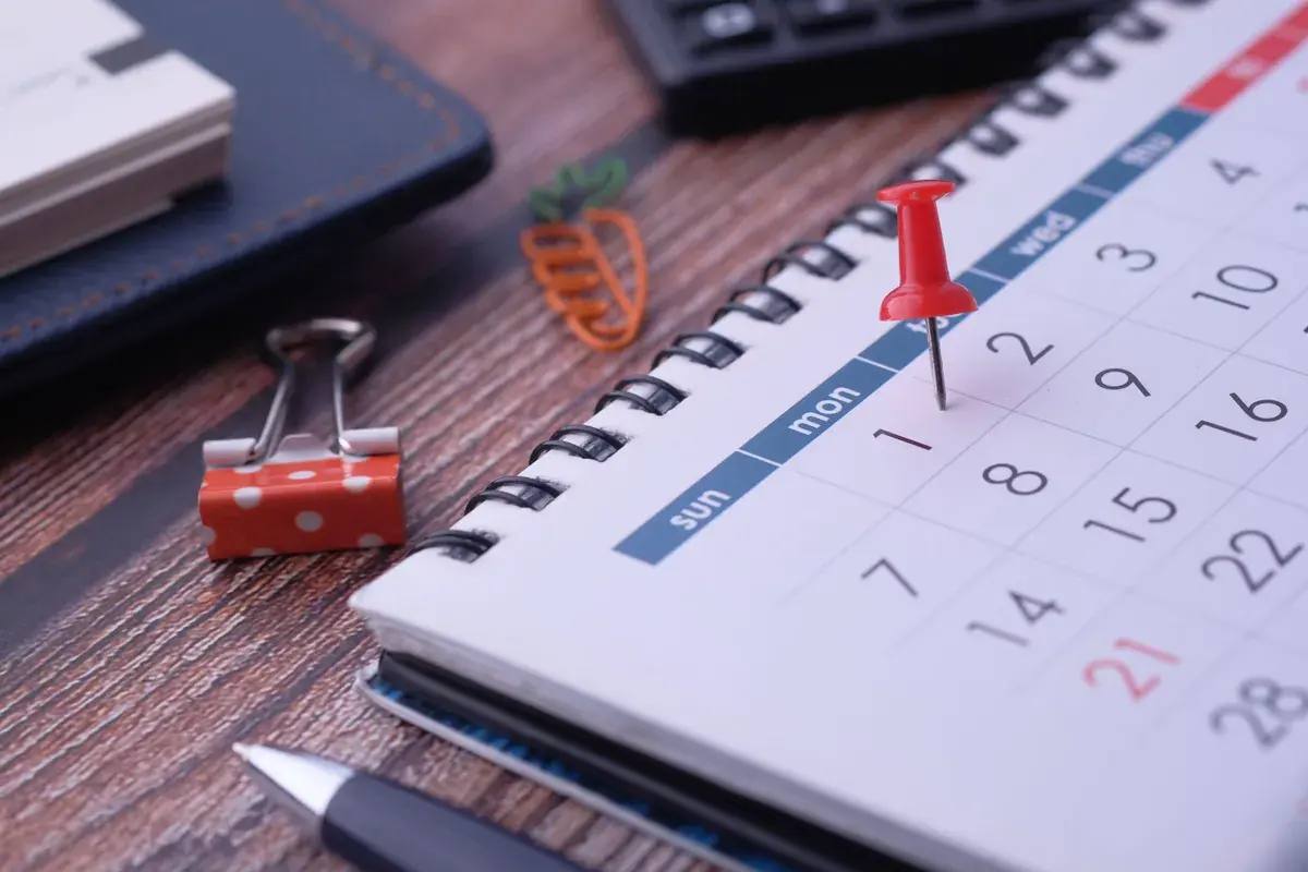 Calendario de escritorio con una tachuela en una fecha, una libreta y una calculadora sobre madera