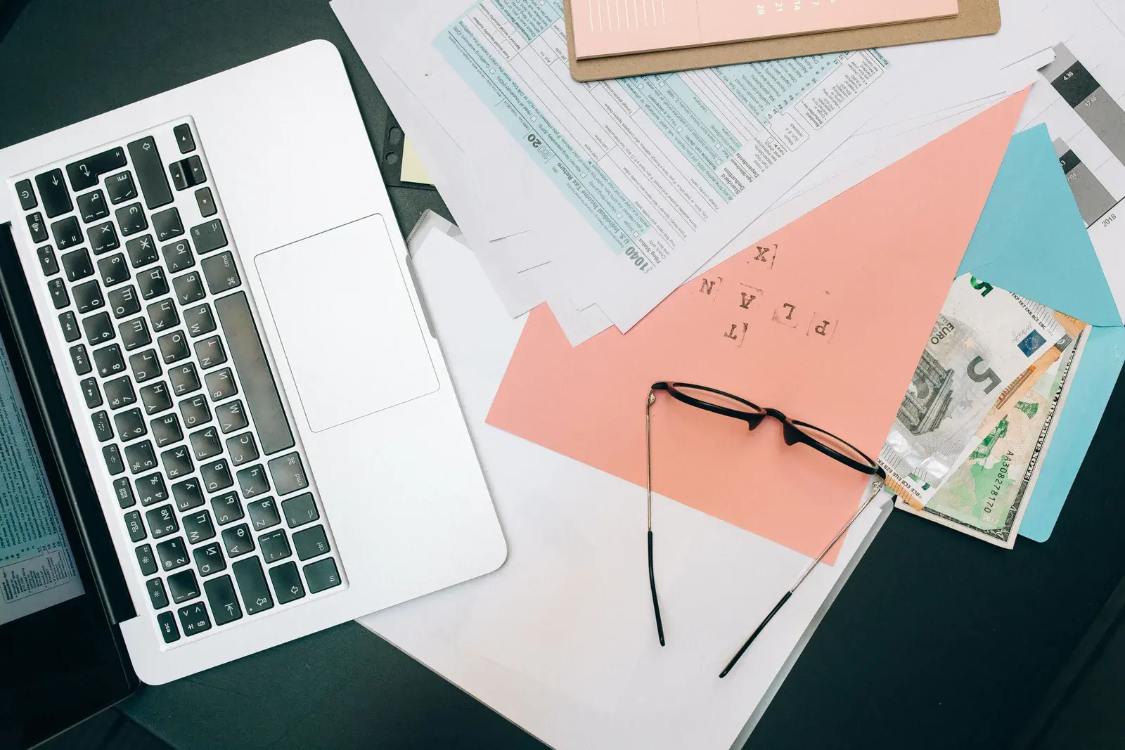 A tidy desk with an open laptop, stacked documents, and eyeglasses under soft window light