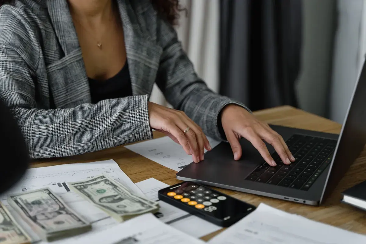 Una mujer trabaja en una laptop con calculadora, efectivo y estados impresos sobre el escritorio