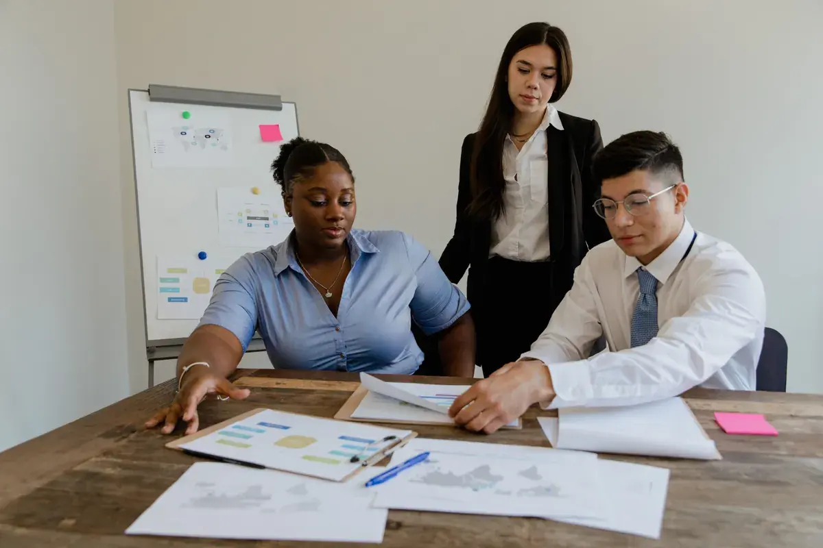 Three colleagues at a wooden table reviewing printed reports and charts beside a whiteboard.