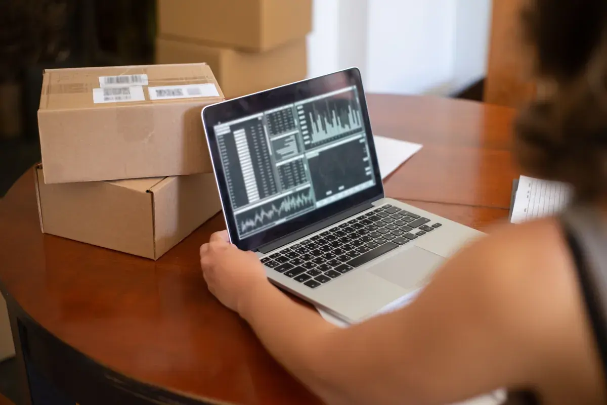 A person works on a laptop showing charts, with labeled shipping boxes stacked on the table beside them.